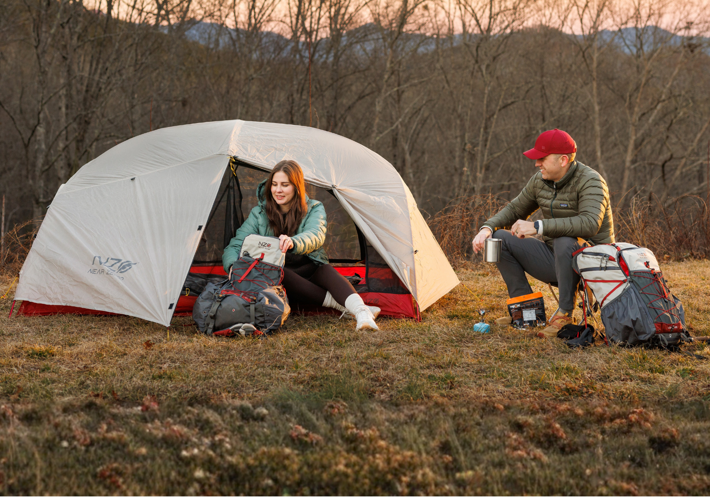 Two people camping outdoors with a tent and backpacks at sunset.
